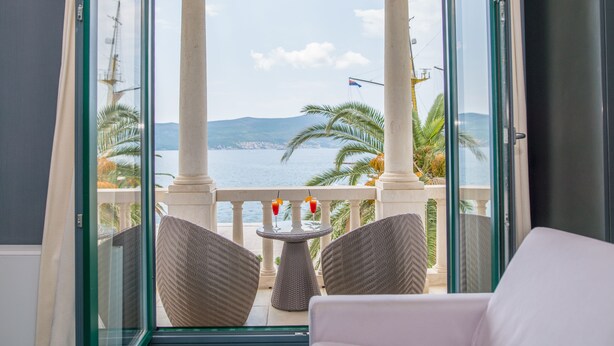 Un balcón con sillas de mimbre, una pequeña mesa redonda con dos bebidas, y vistas al mar y a las montañas.