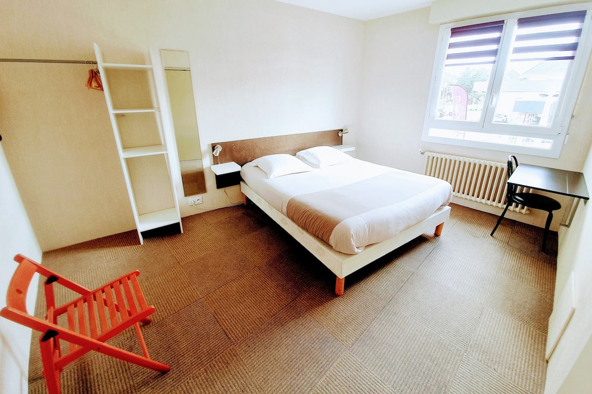 A hotel room with a bed, a desk, a chair, a radiator, a window, and a wardrobe.