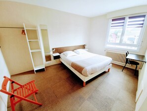 Habitación de hotel con cama, escritorio, silla, radiador, ventana y armario.