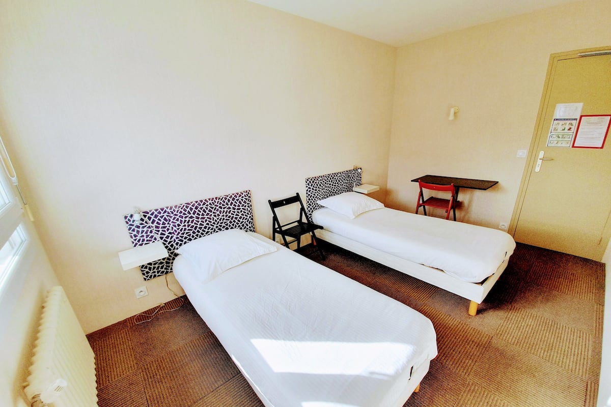A room with two single beds, a small table, a chair, and a radiator.