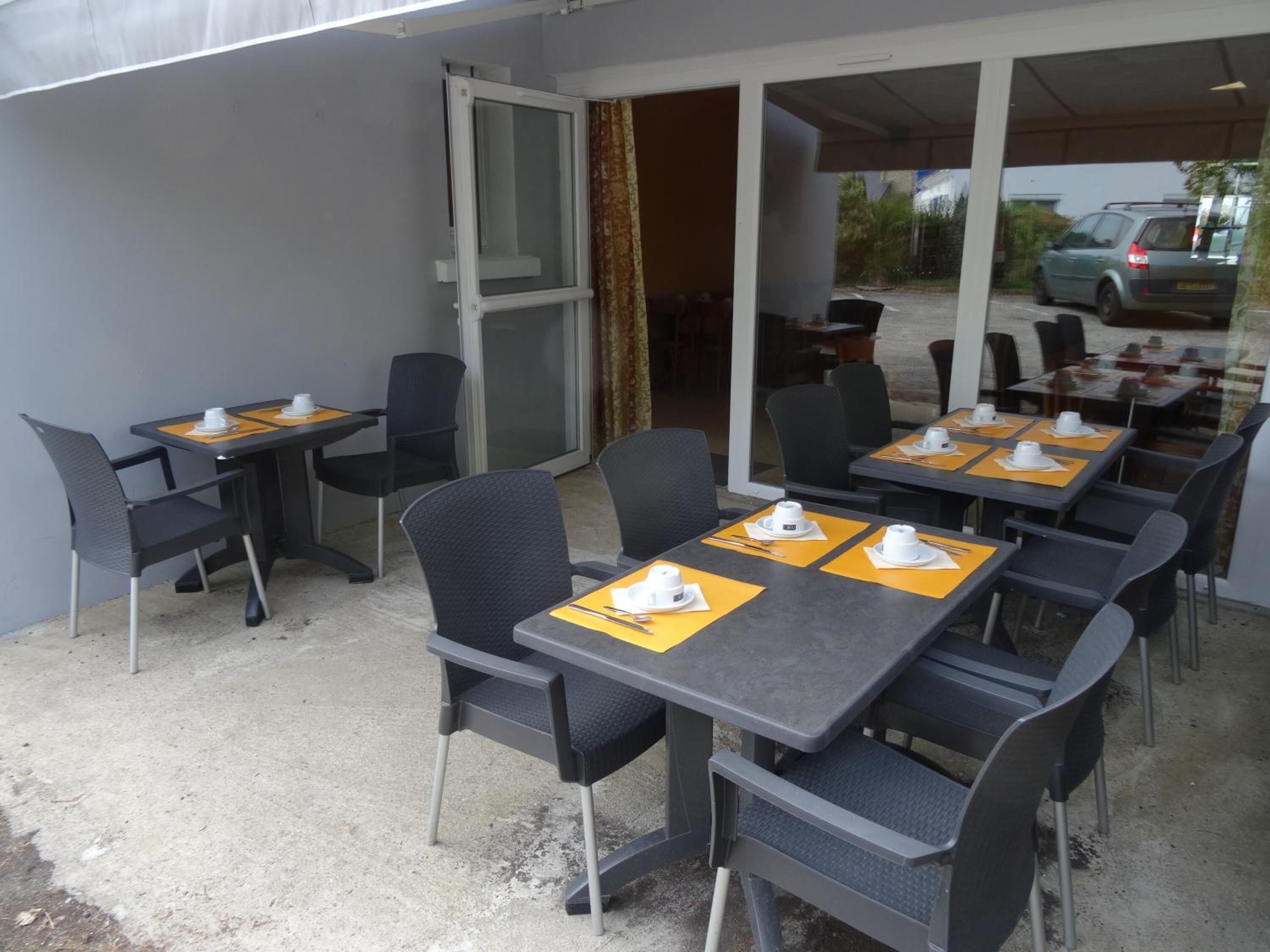 Een terras met tafels en stoelen, een glazen deur en zicht op geparkeerde auto's.
