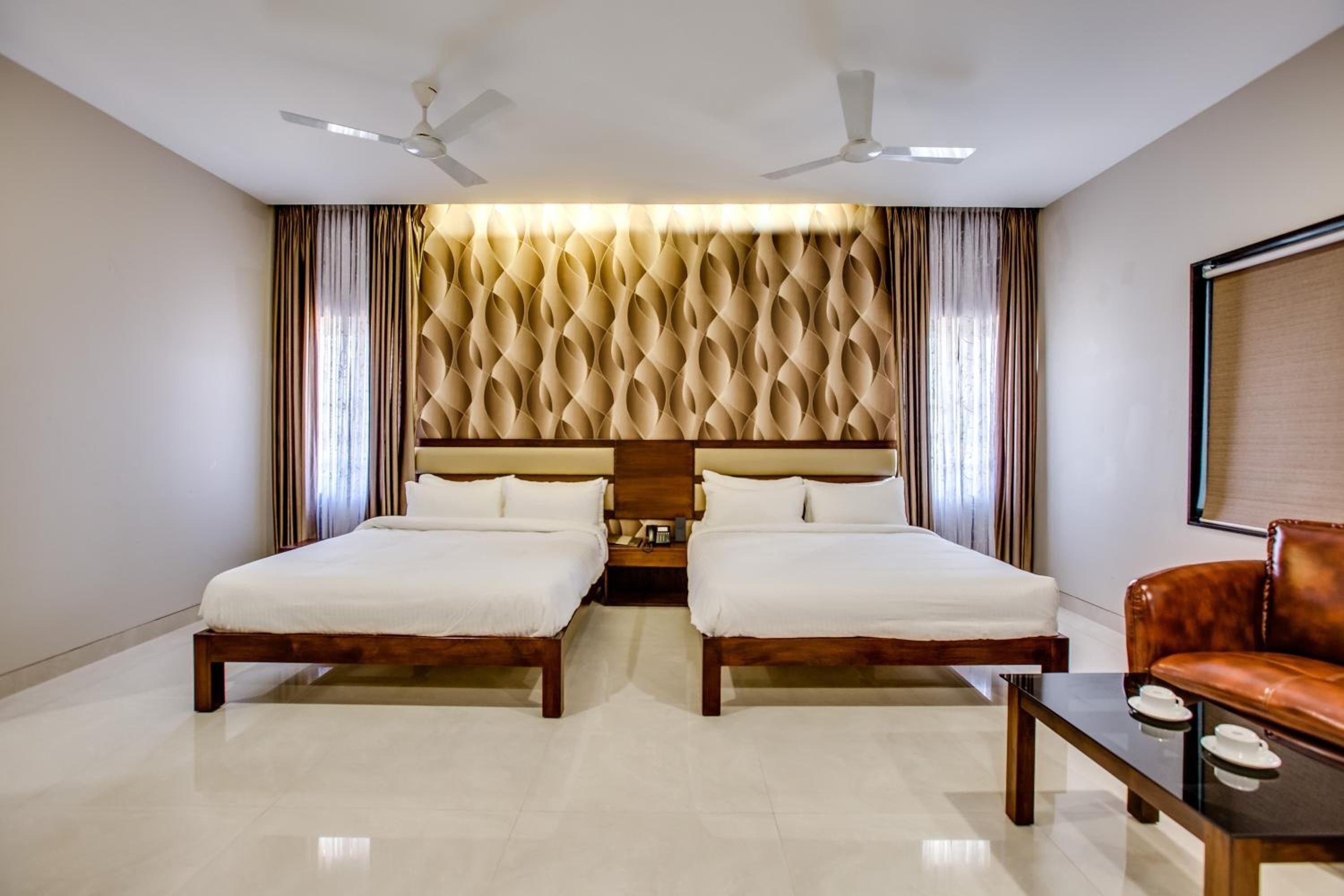 Habitación de hotel con dos camas, cabecera de madera, ventilador de techo, sofá de cuero y una pequeña mesa.