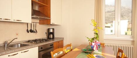 A compact kitchen with white cabinets, a gas stove, and a coffee maker. A dining table with chairs is set up near a window with a view of a hill.