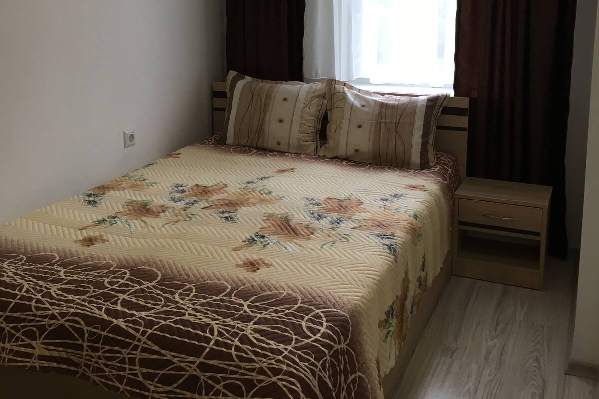 A bedroom with a bed, a nightstand, a window with curtains, and a wall socket.