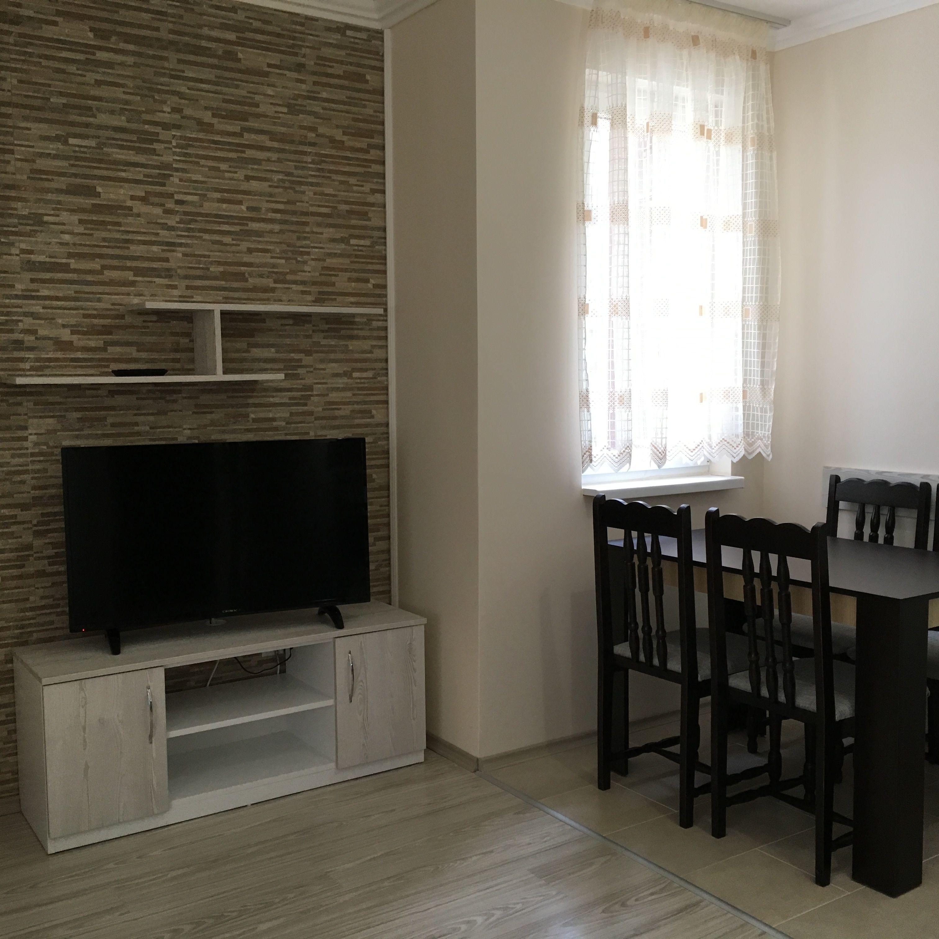 A living room with a flat-screen TV on a wooden stand, a dining table with chairs, and a window with lace curtains.