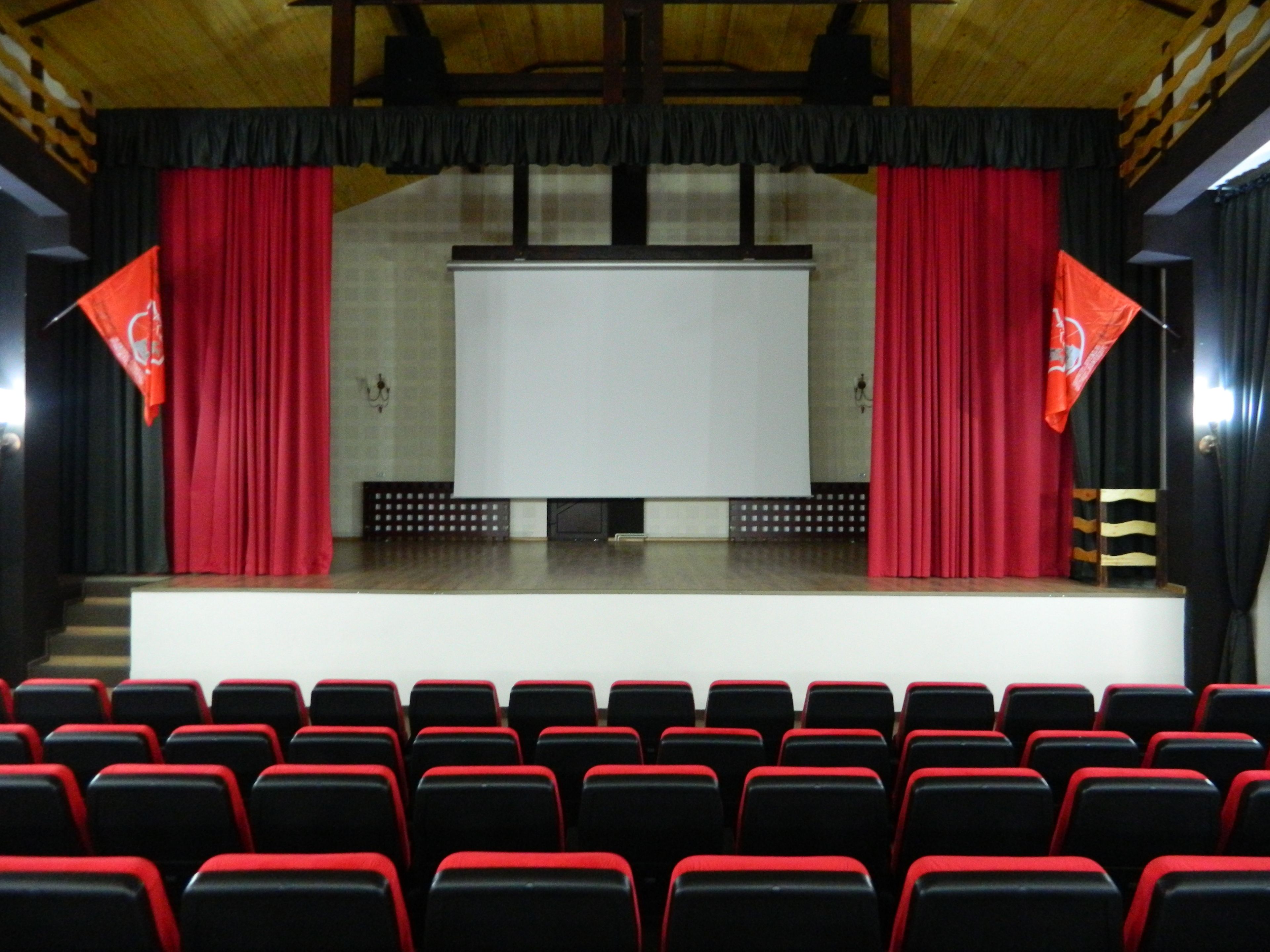 An auditorium with rows of red and black seats, a stage with a white screen, and red curtains.