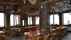 Un comedor rústico con mesas y sillas de madera, pilares de piedra y ventanas amplias.