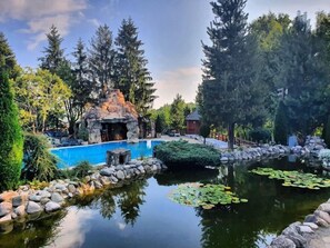 A serene outdoor pool area with a natural rock formation, surrounded by lush greenery and tall trees. - Club Vila Bran (Bran)