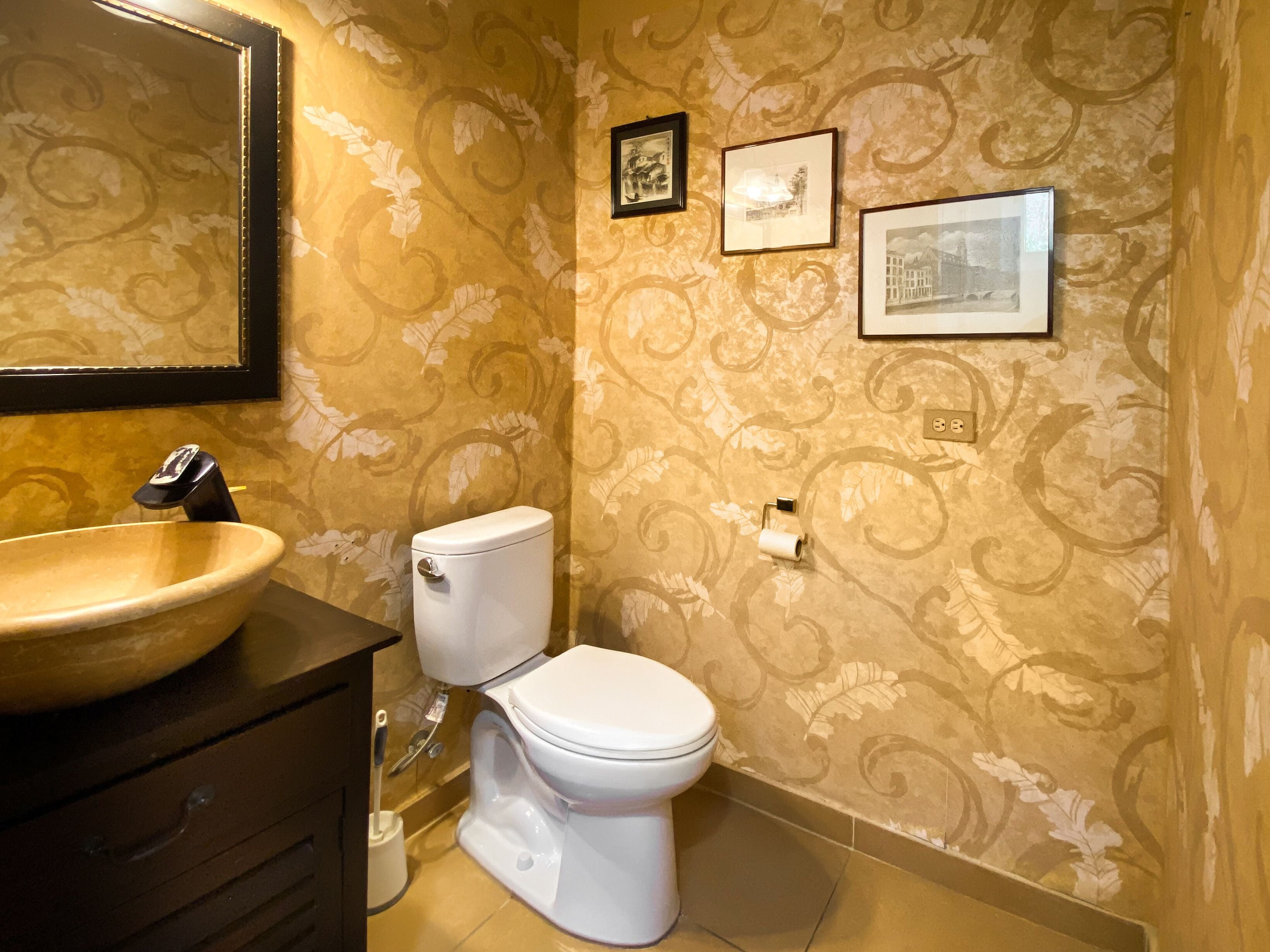 Ein Badezimmer mit einer beige-gemusterten Wand, einem hölzernen Waschtisch mit großer Keramikwaschbecken, einer weißen Toilette und gerahmten Bildern an der Wand.