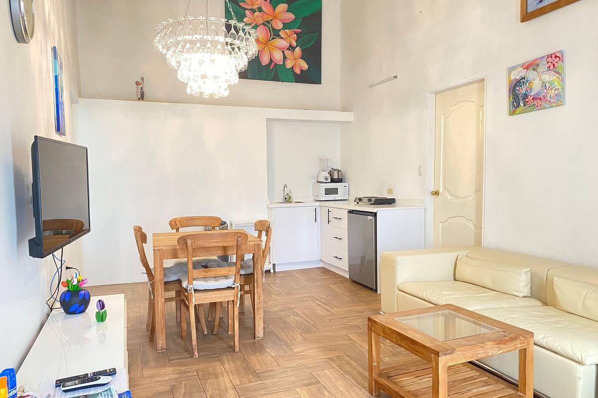 Un espace de vie compact comprenant un coin repas, un canapé, une table basse, un téléviseur, une cuisine et un lustre.