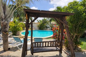 A wooden pergola with a bench, a pool, and surrounding tropical plants. - Casalina Garden Apartment Aruba (Noord)