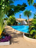 A swimming pool with a thatched-roof structure, surrounded by palm trees and a brick pathway.