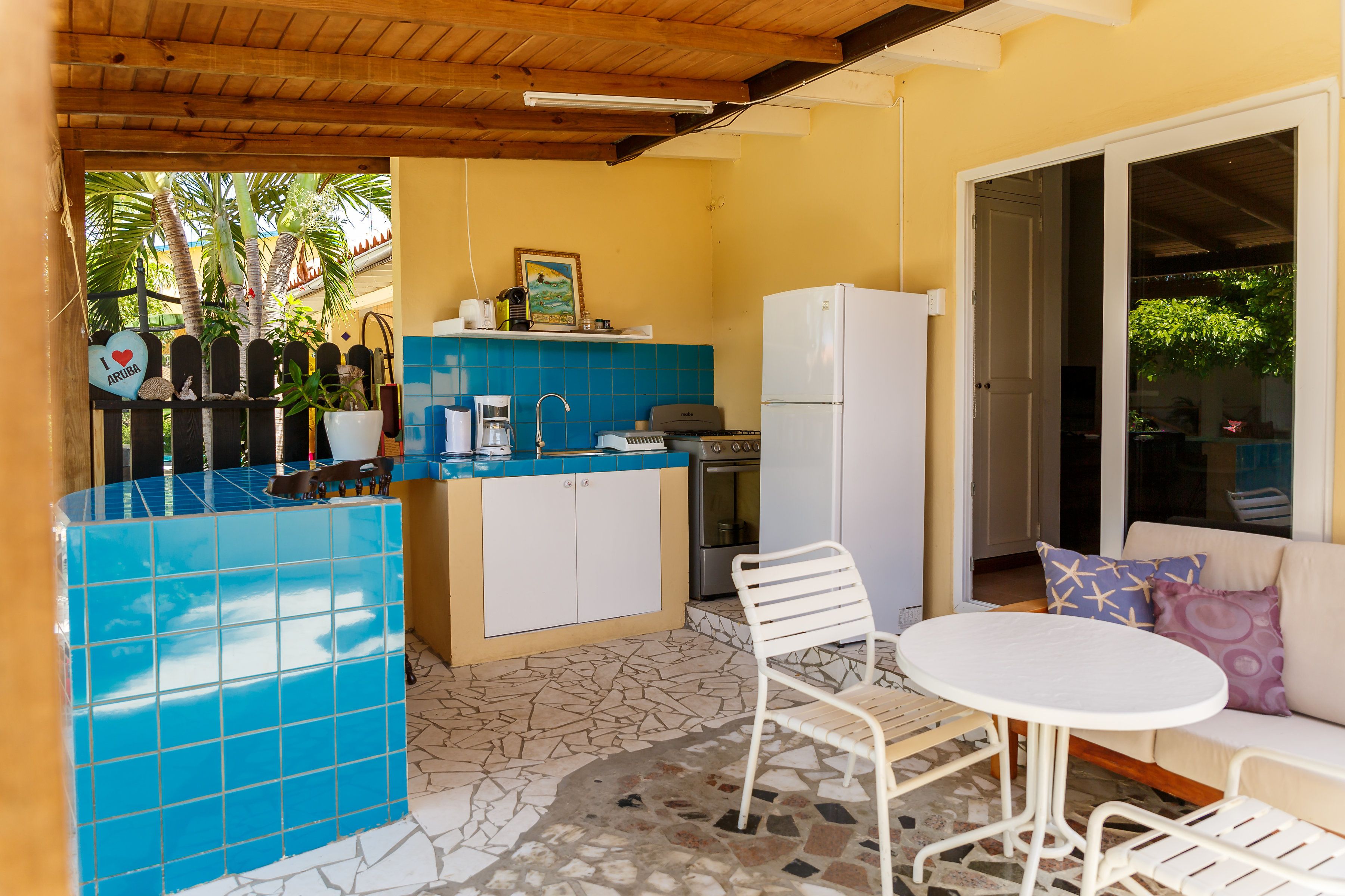A compact kitchen with a blue tiled backsplash, a white refrigerator, and a small dining area with a white table and metal chairs.