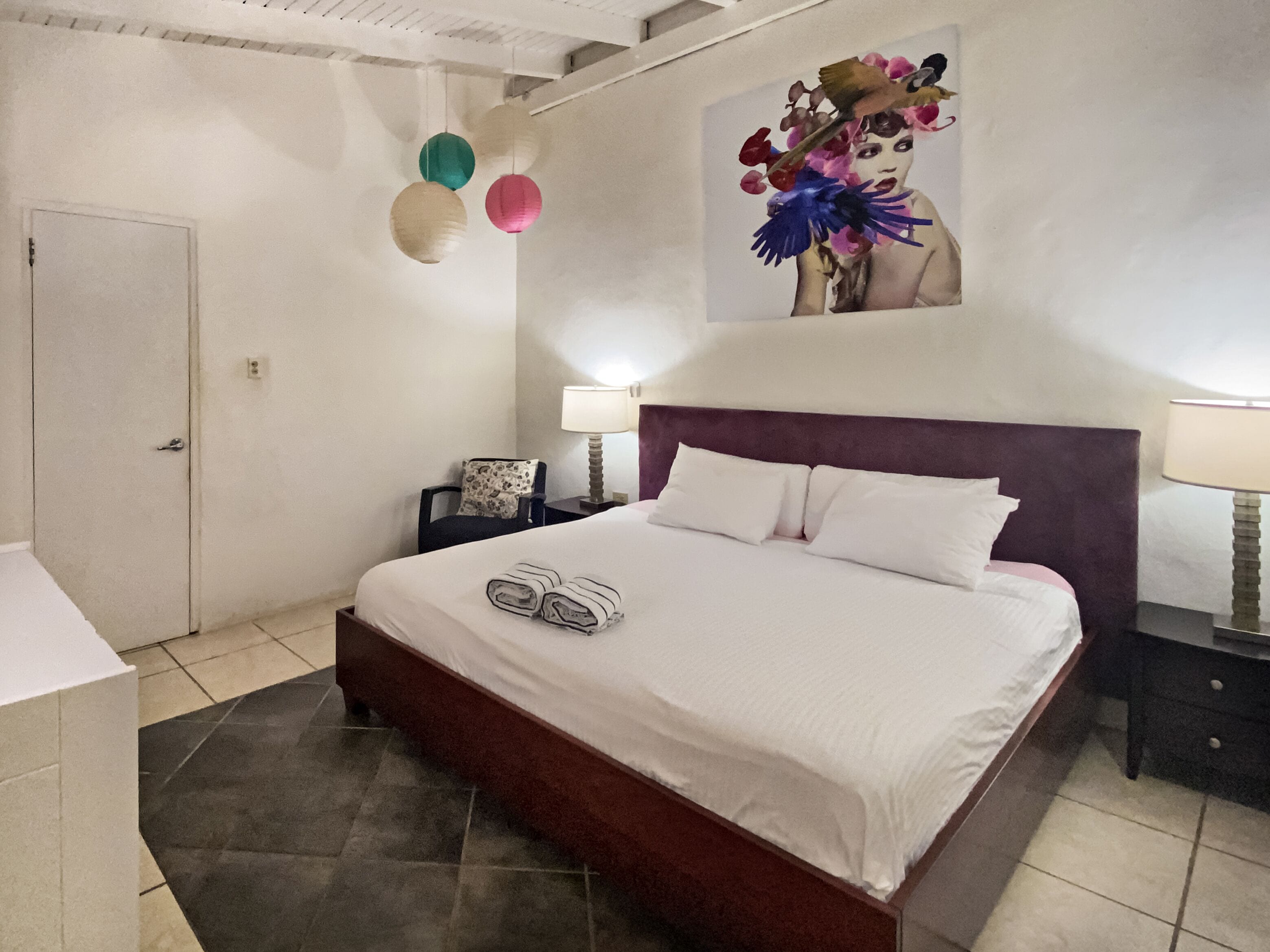 Un dormitorio con una cama grande, una mesita de noche, una silla, una puerta y una pared con un cuadro.