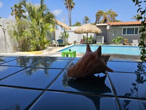 A pool area with a clear blue pool, lounge chairs, and a thatched umbrella. - Casalina Garden Apartment Aruba (Noord)