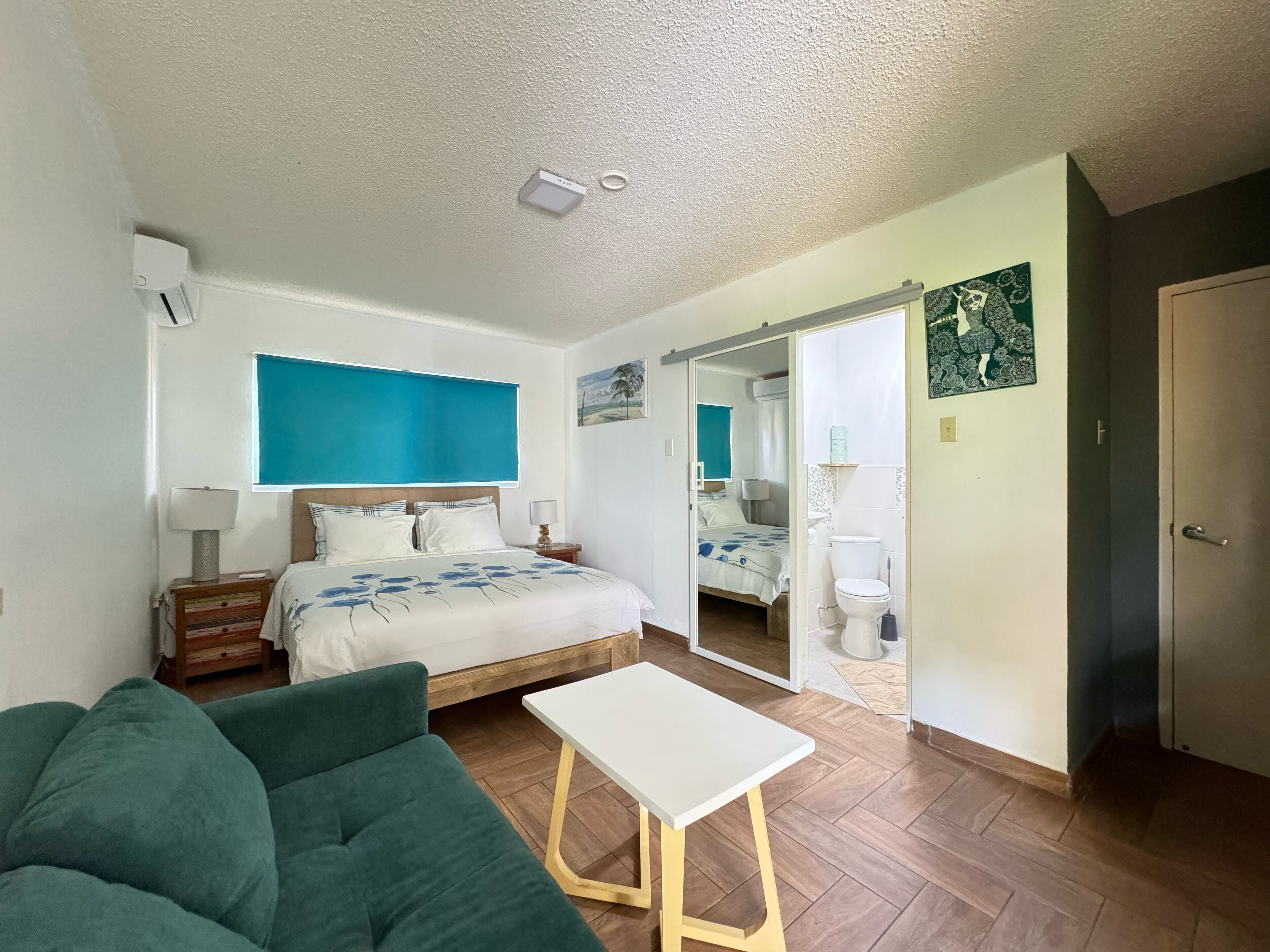 A bedroom with a bed, bedside tables, a green sofa, a small white table, and a bathroom visible through an open door.
