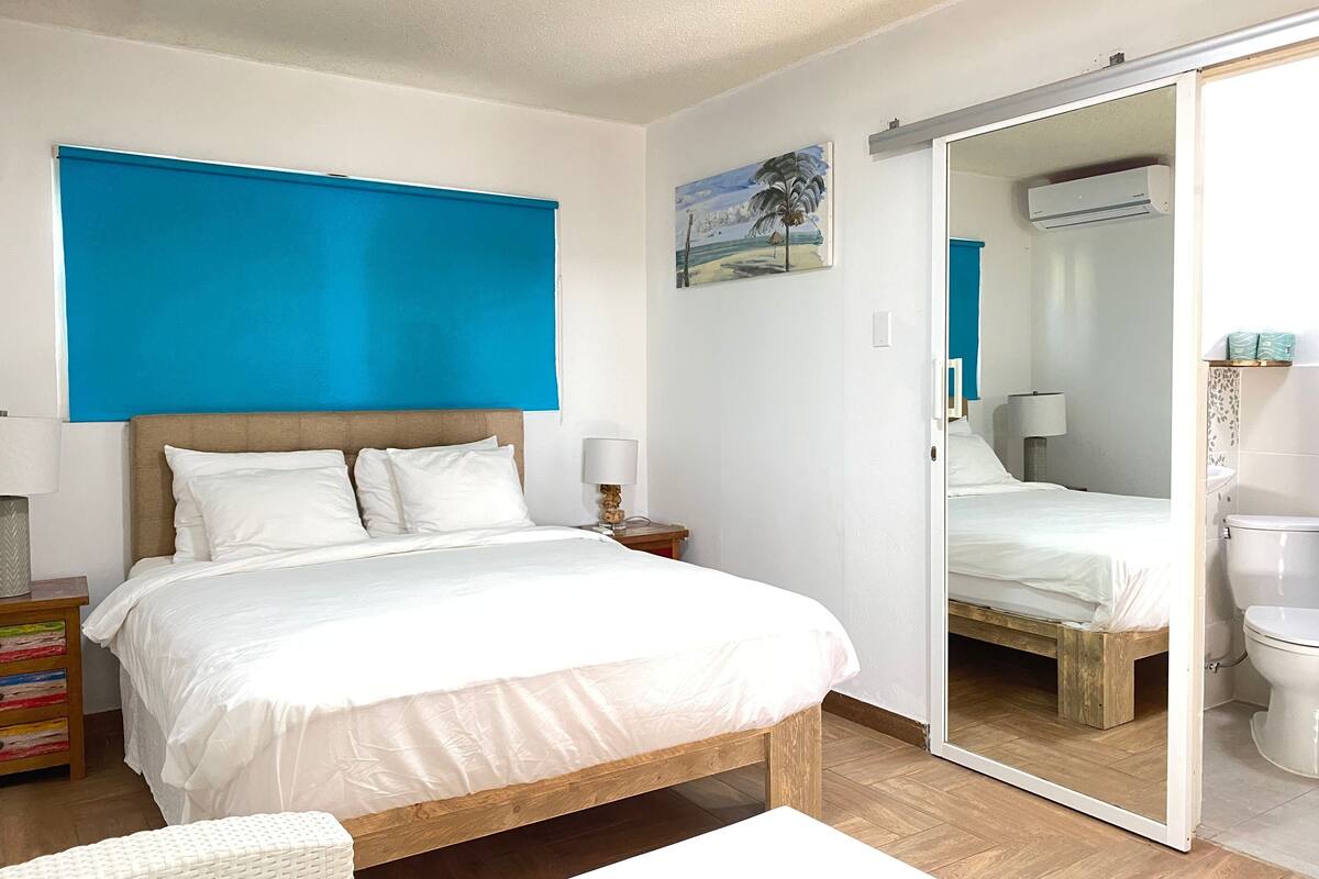 Une chambre à coucher avec un lit, une table de chevet, une lampe, un banc en bois et une salle de bain visible à travers une porte ouverte.
