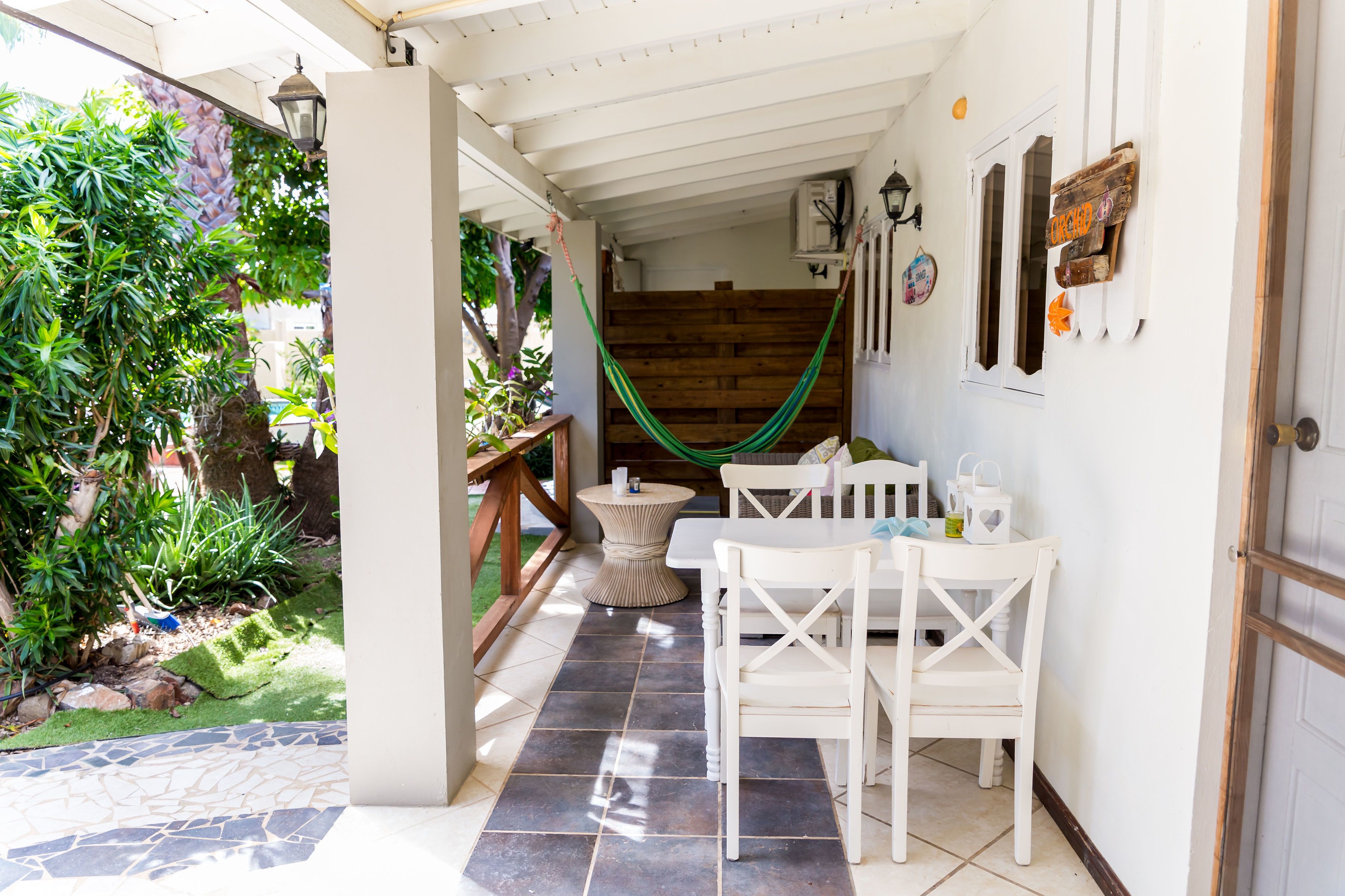 Un patio cubierto con un banco de madera, una mesa blanca con sillas y una hamaca.