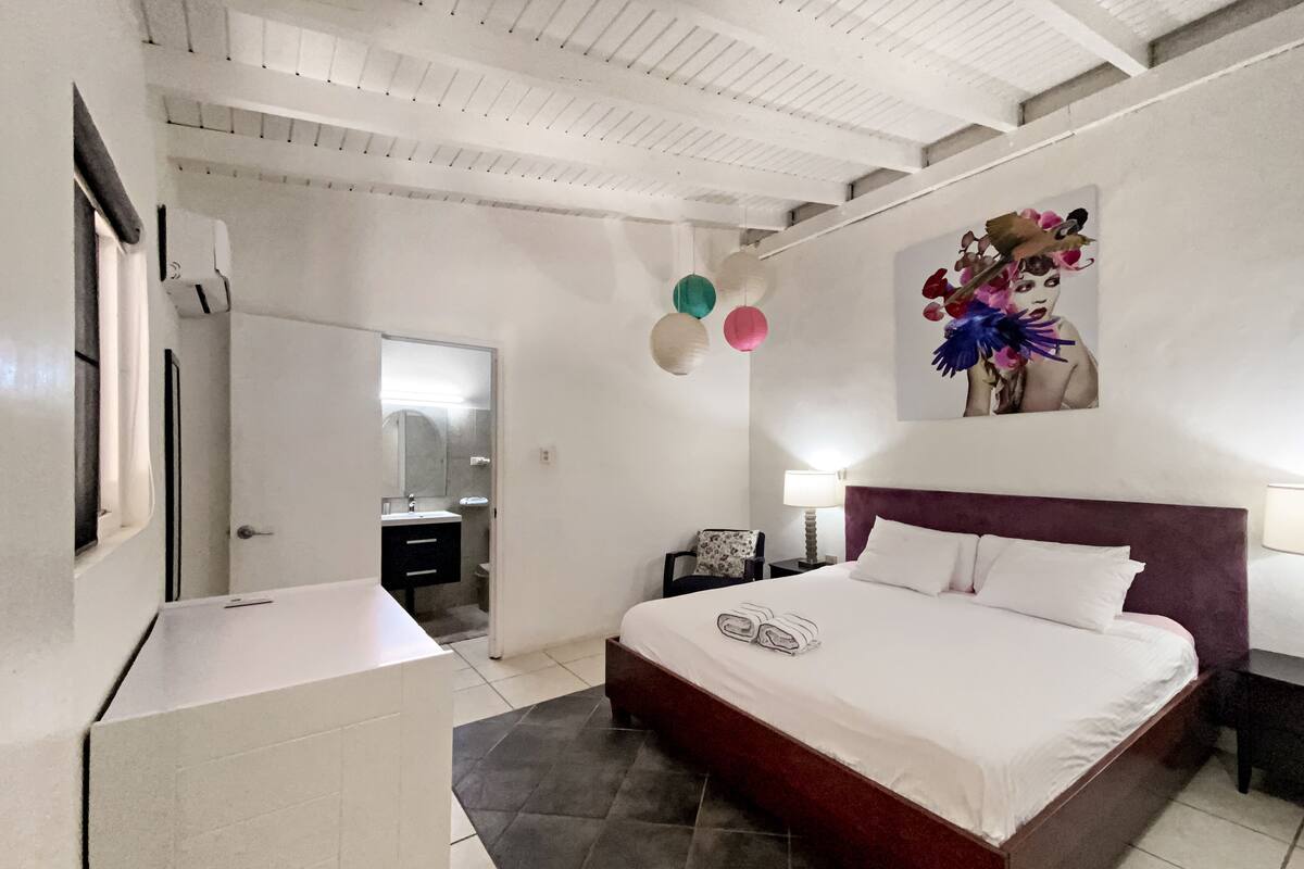 Une chambre moderne avec un grand lit, une table de chevet, une commode, un miroir et une salle de bain visible à travers une porte ouverte.