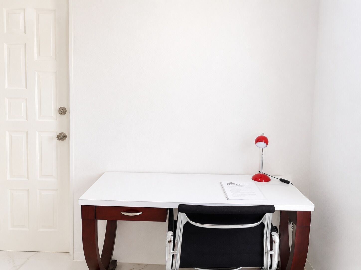 A white desk with a red lamp, a black chair, and a door in the background.