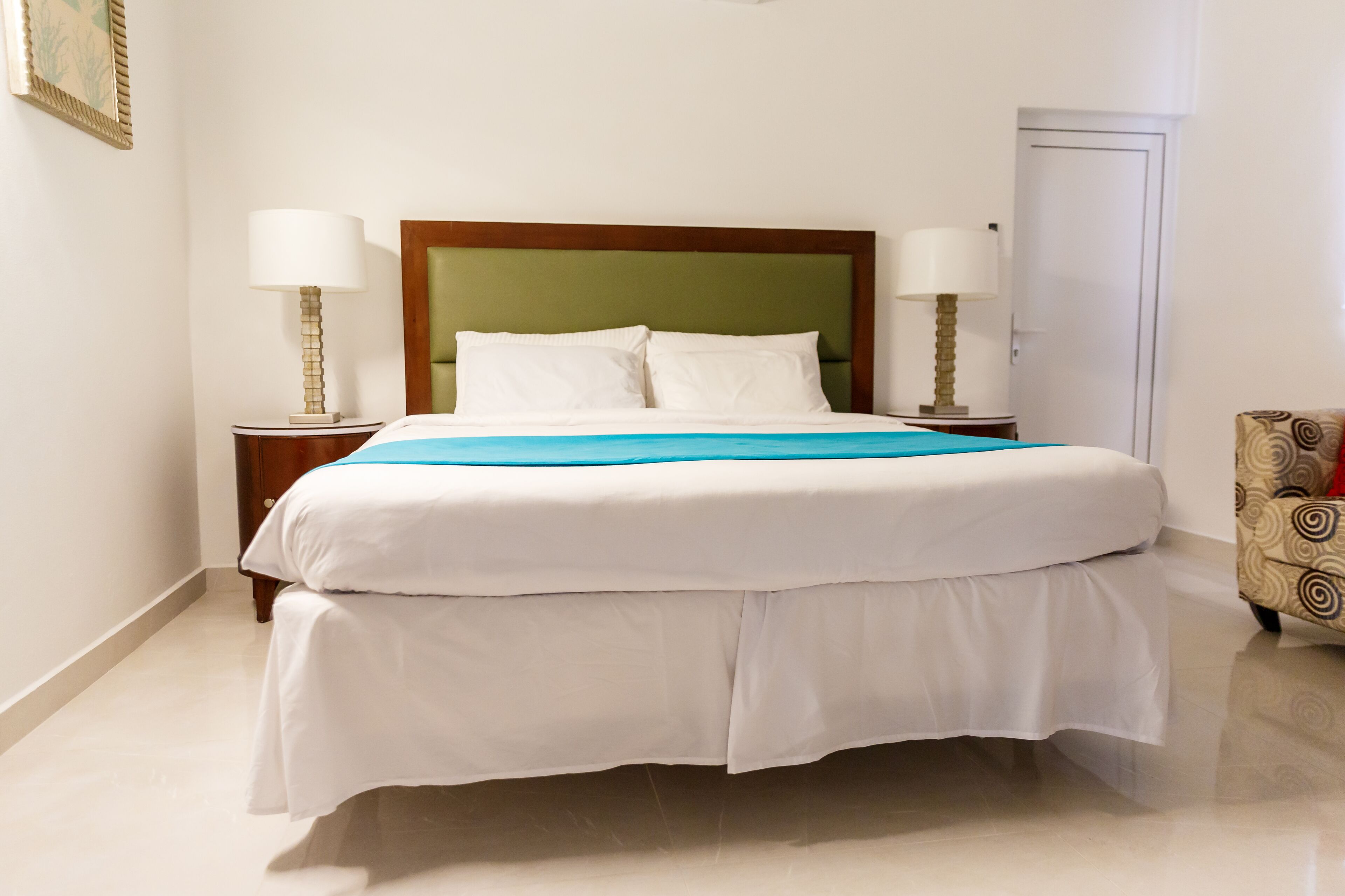 A neatly made bed with a green headboard, two bedside lamps, and a patterned armchair.