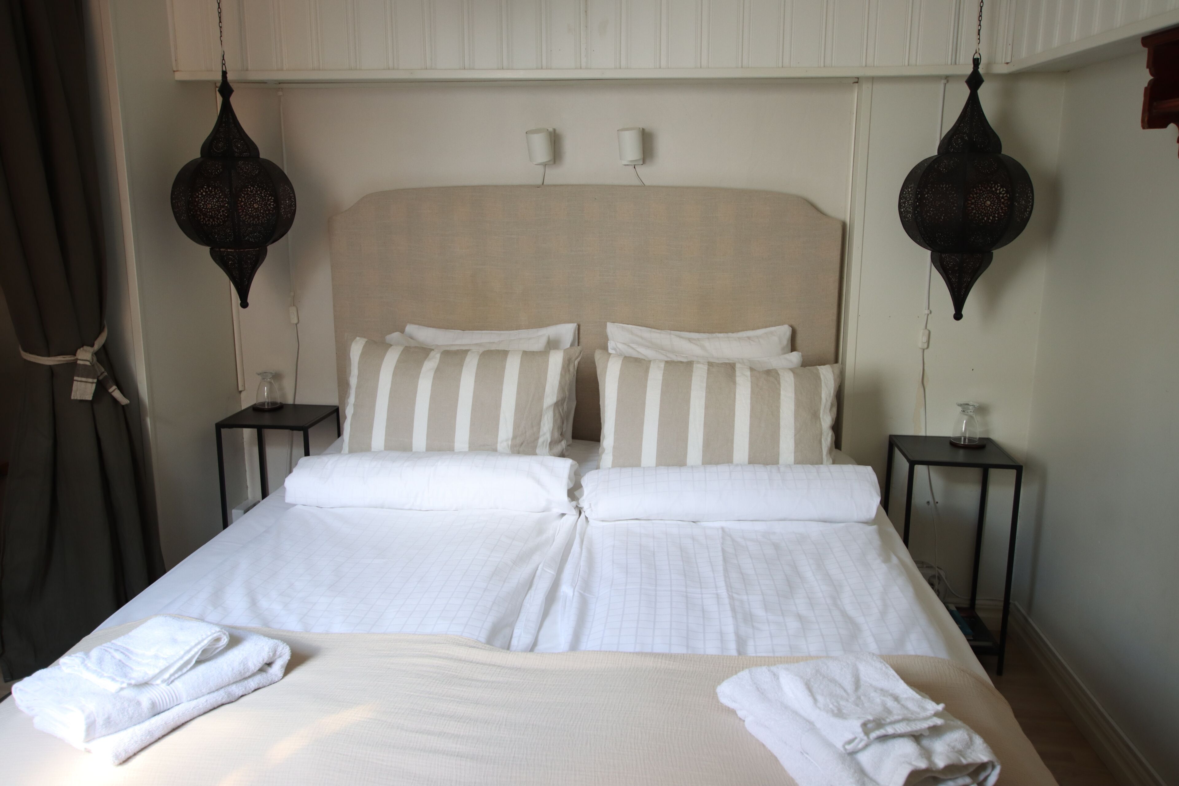 Una cama doble con ropa de cama blanca y almohadas a rayas, dos mesitas de noche con lámparas y una lámpara colgante.
