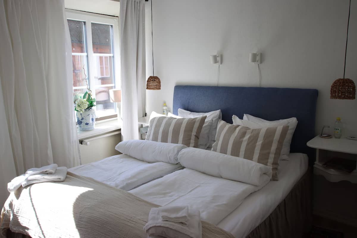 A hotel room with a bed, a window with curtains, a bedside table with a lamp, and a wall-mounted light fixture.