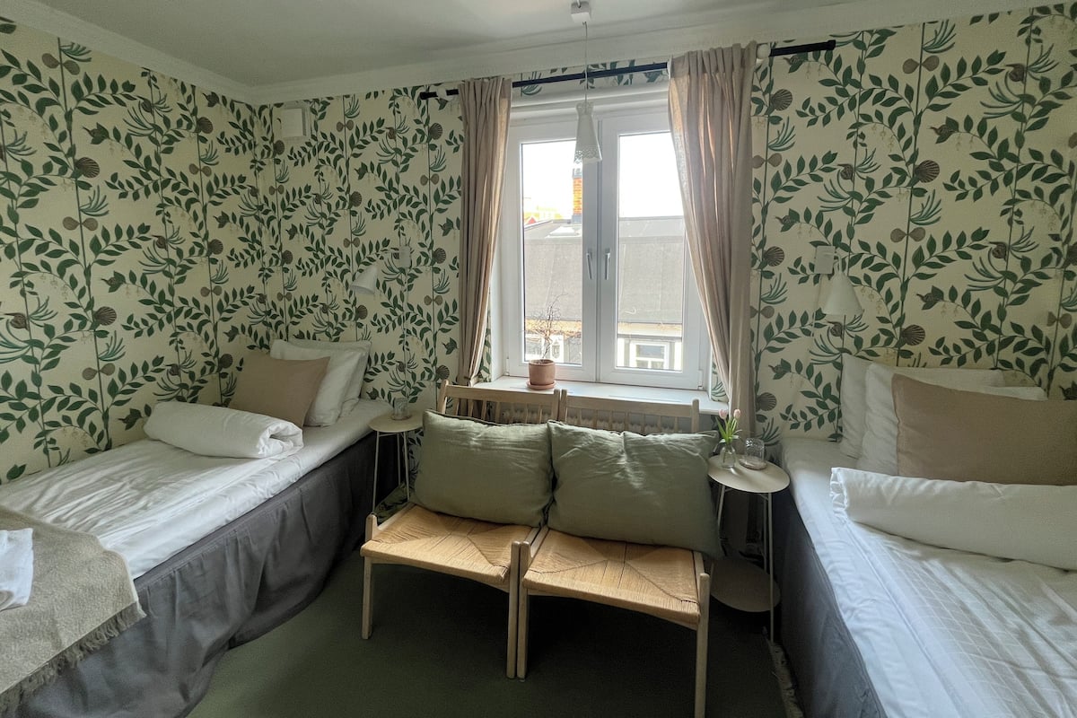 A room with two single beds, a small table, a bench, and a window with curtains.