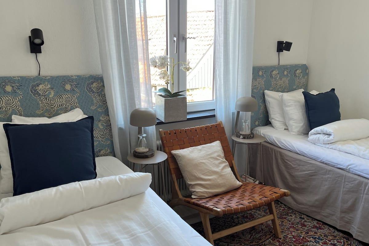 A hotel room with two beds, a chair, a small table with a lamp, and a window with curtains.