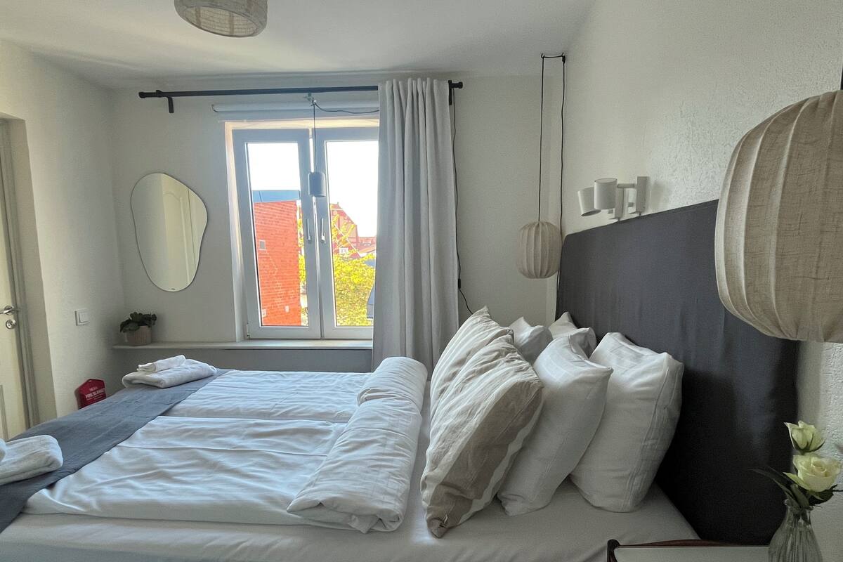 A neatly made bed with white linens, a bedside table with a vase of flowers, a large window with curtains, and a wall-mounted mirror.