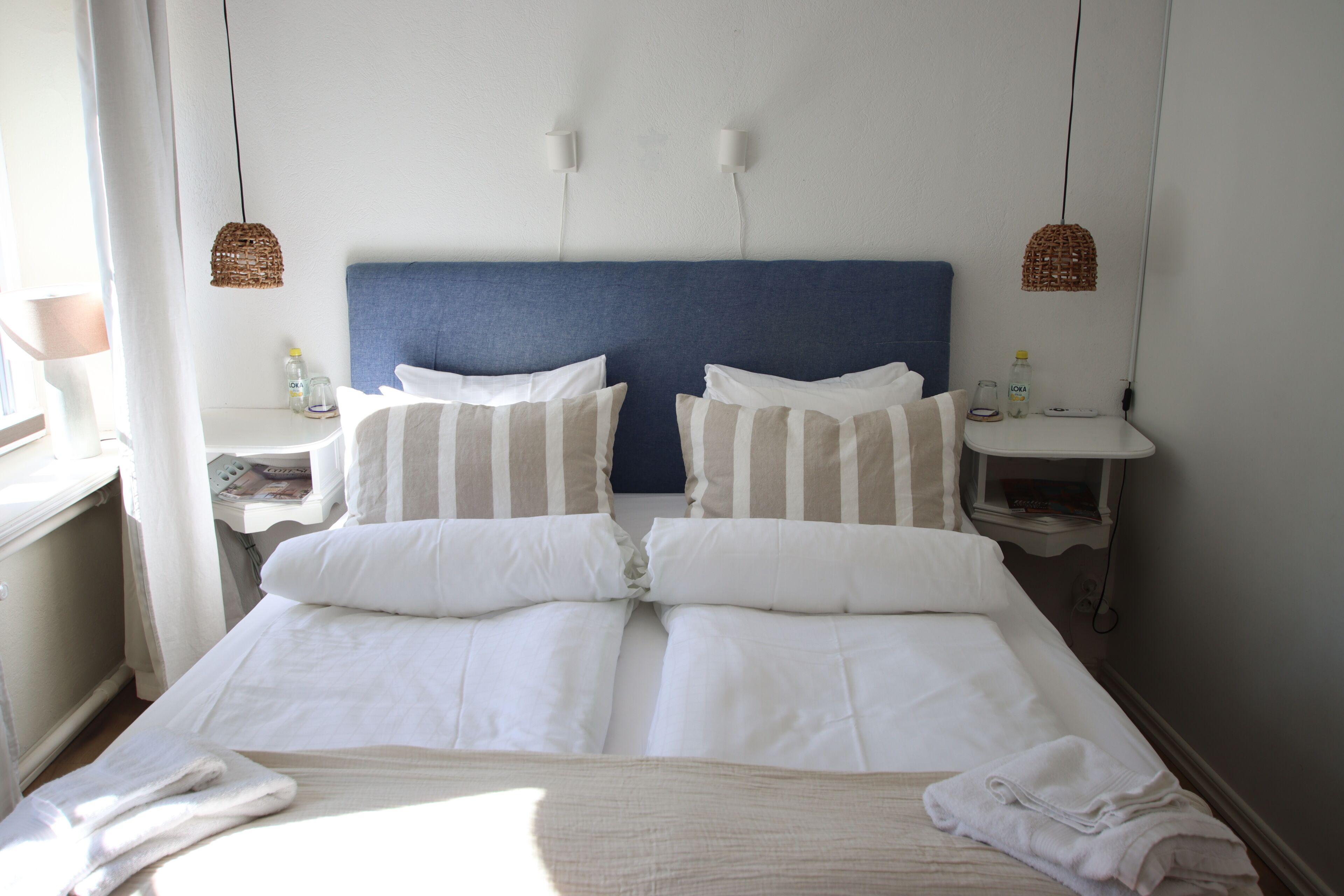Een tweepersoonsbed met witte en gestreepte kussens, een blauw hoofdbord, twee nachtkastjes met lampen, en een raam met gordijnen.