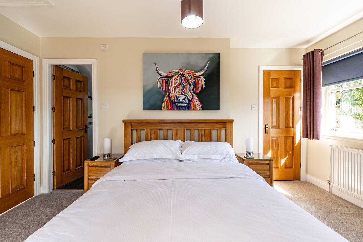 A bedroom with a large bed, wooden headboard, two bedside tables with lamps, a painting of a bull's head on the wall, and a window with curtains.