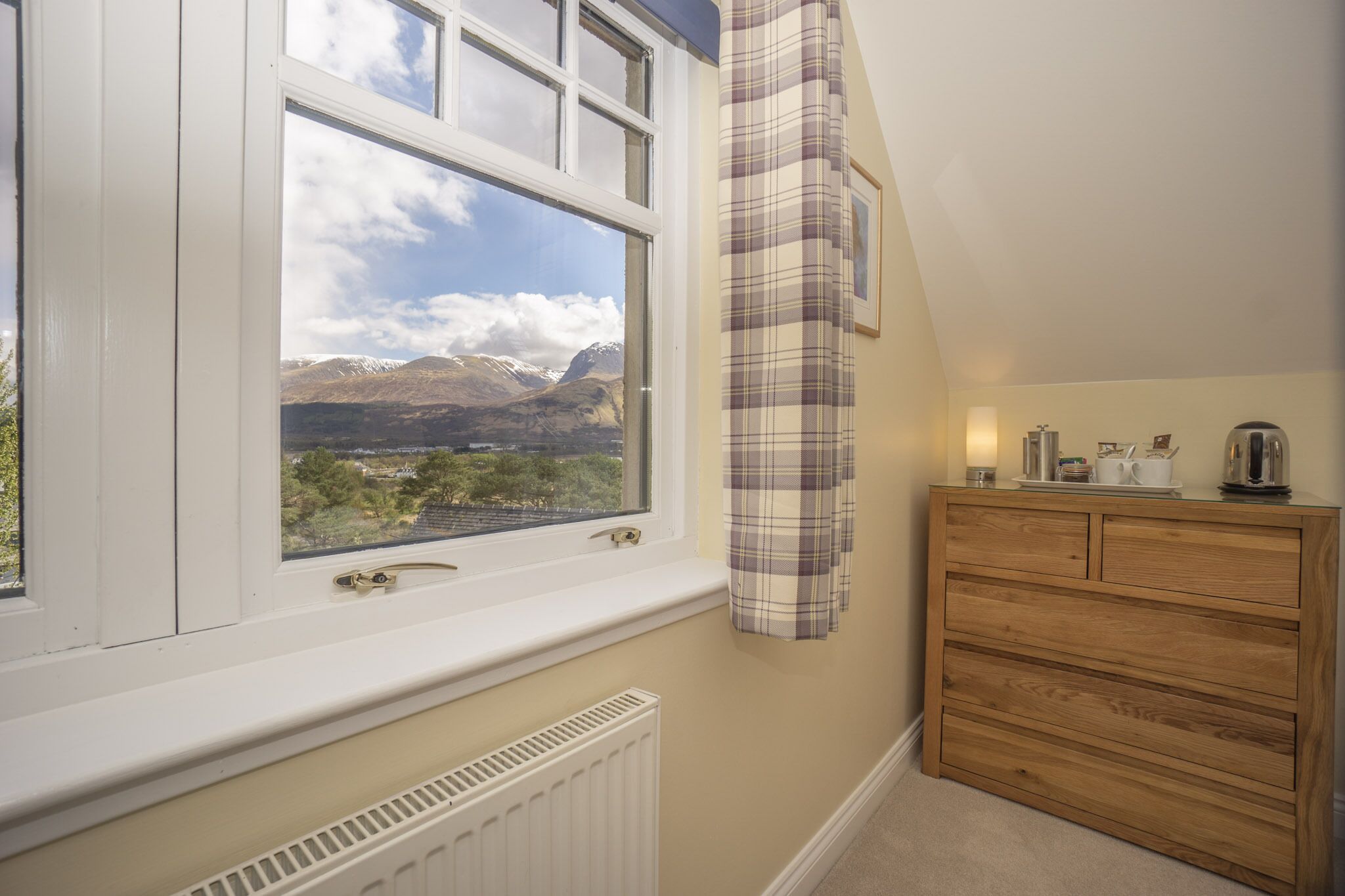 A room with a window overlooking a mountainous landscape, a plaid curtain, and a wooden dresser with a kettle on top.