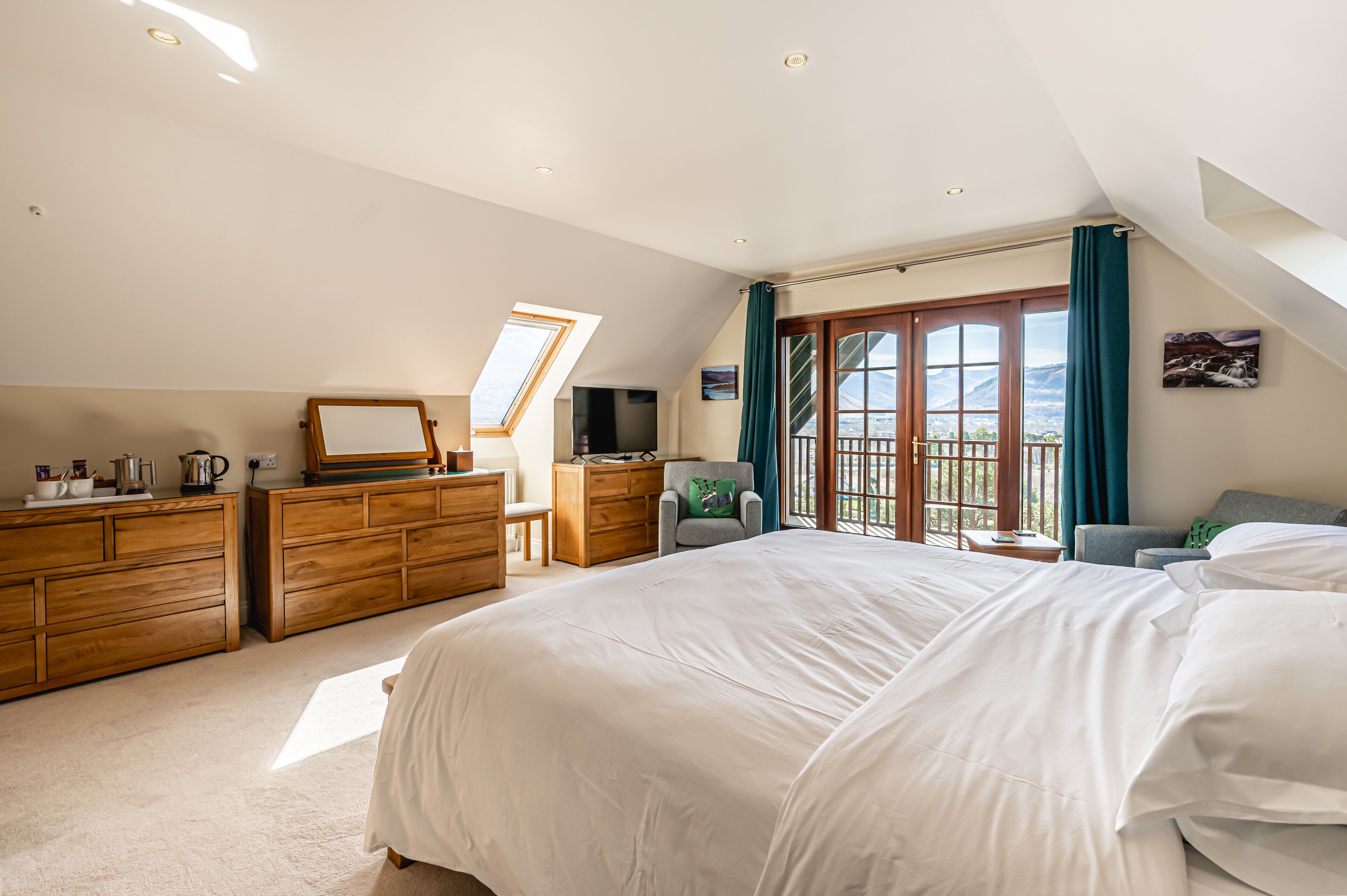 A bedroom with a large bed, wooden dresser, desk, and a view of the outdoors through a large window.