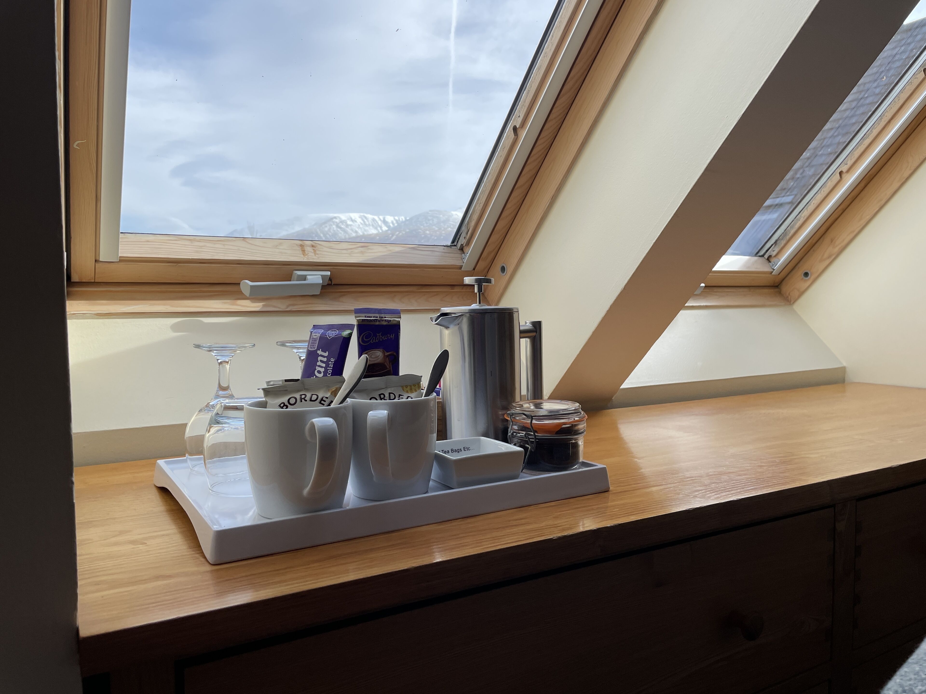 Ontbijtarrangeement met koffiekopjes, een theepot en een potje op een houten tafel bij een dakraam.