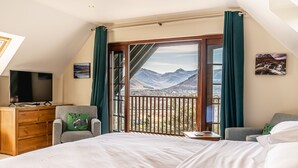 A bedroom with a large bed, a wooden dresser, a TV, and a view of mountains through a sliding door. - Treetops B&B (Fort William)