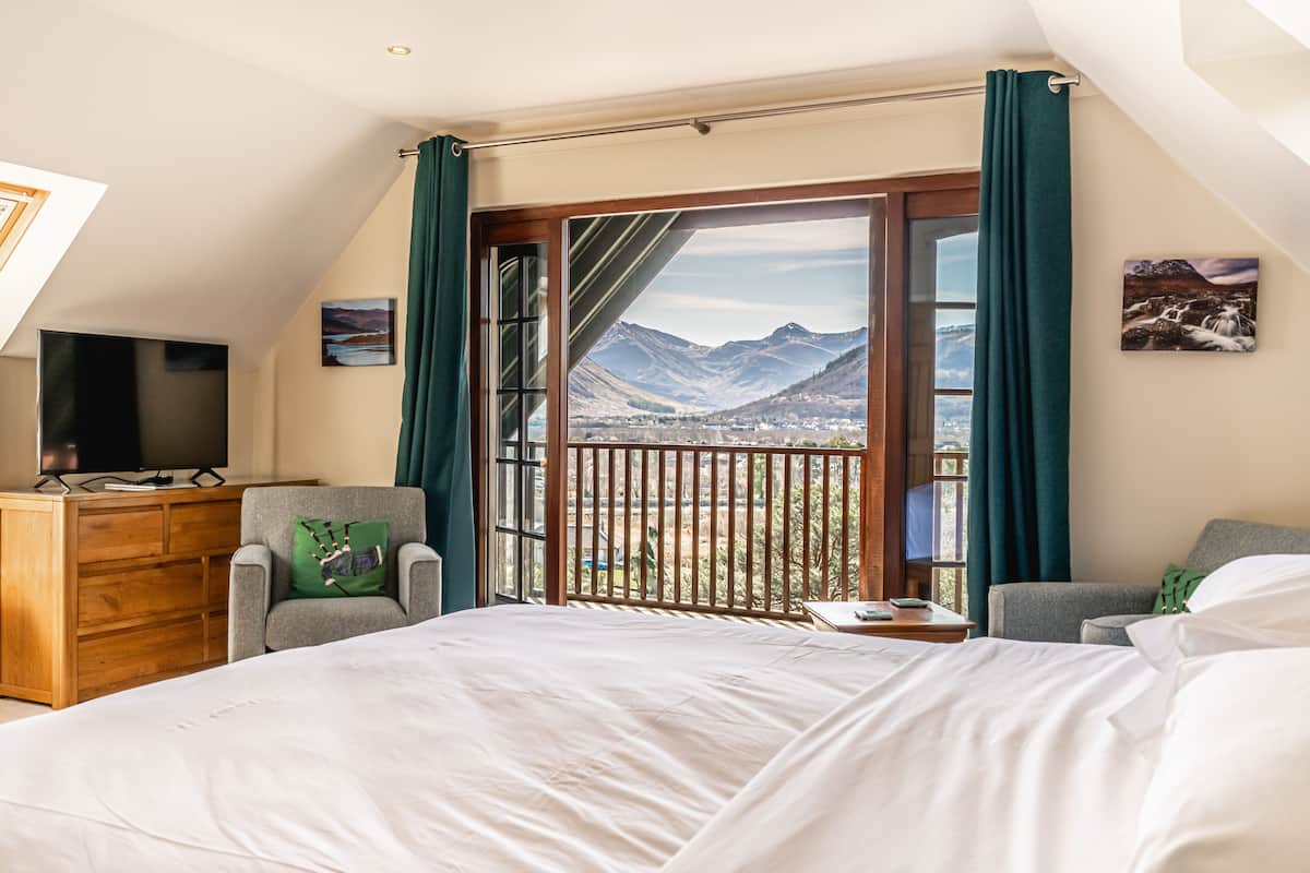 A bedroom with a large bed, a wooden dresser, a TV, and a view of mountains through a sliding door.