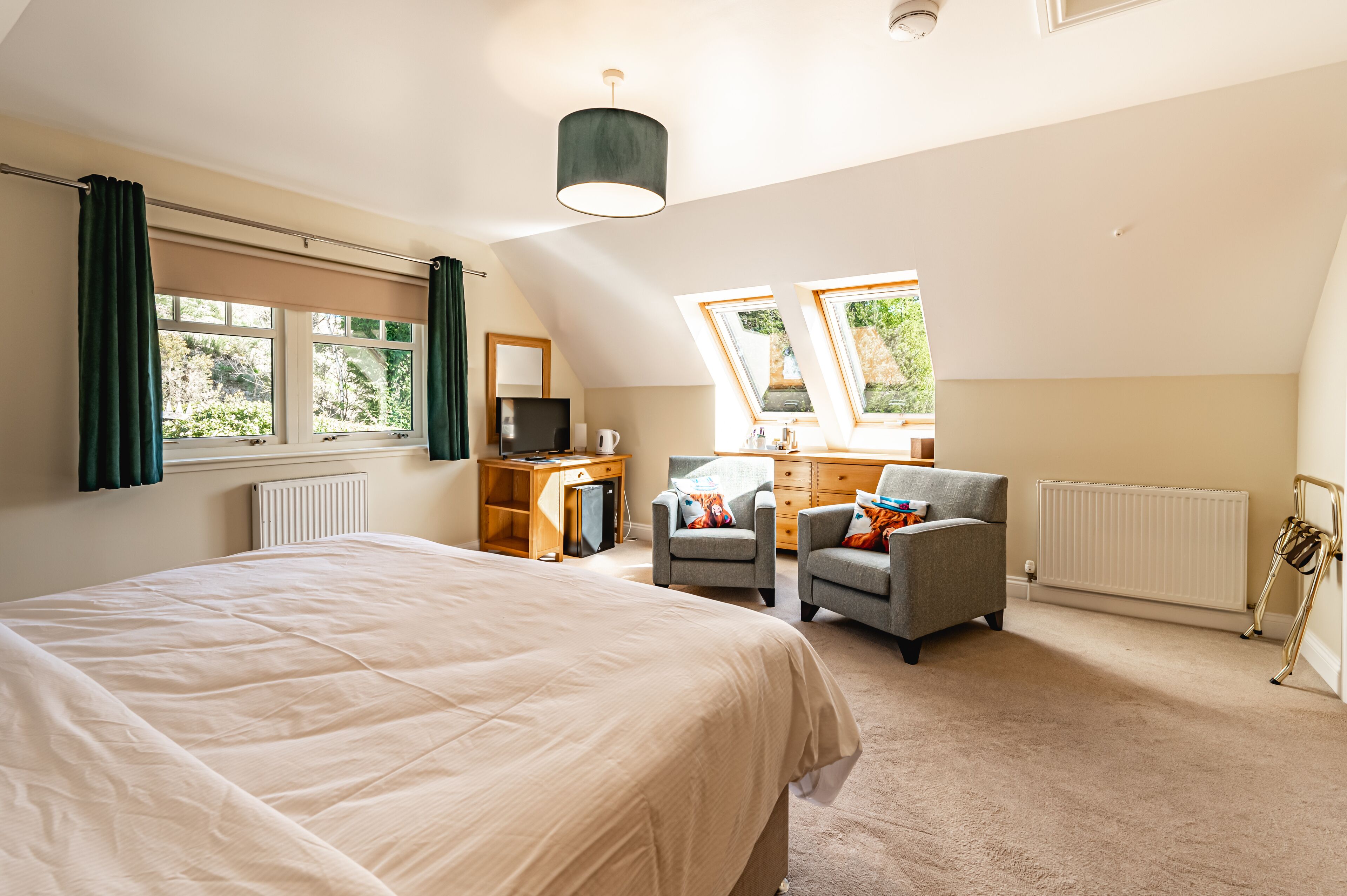 Une chambre à coucher avec un grand lit, deux fauteuils, un bureau, une télévision et une fenêtre donnant sur un paysage verdoyant.