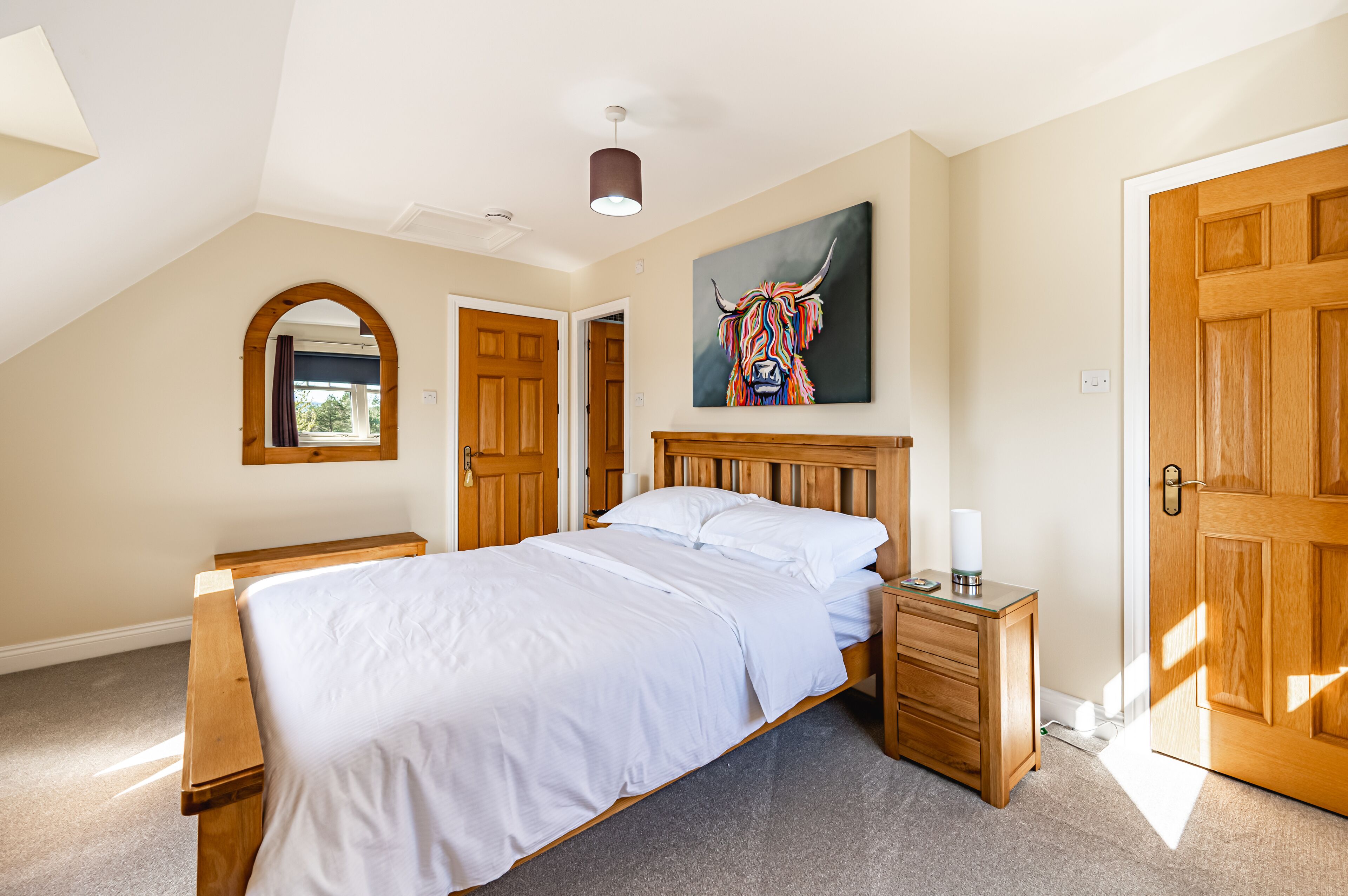 A bedroom with a wooden bed, a nightstand, a wooden dresser, a large window with curtains, and a ceiling light.