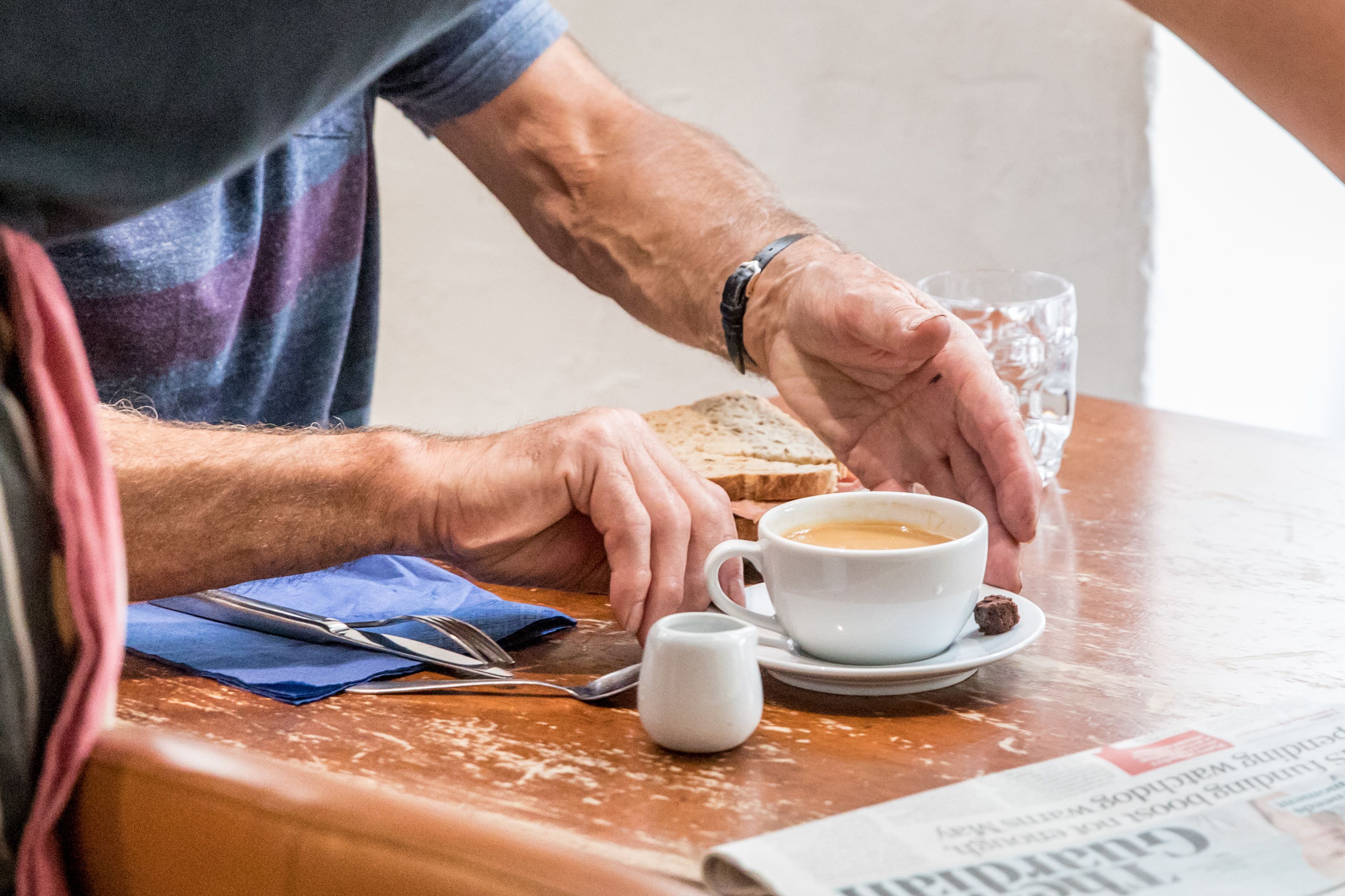 En person sitter vid ett bord med en kopp kaffe, en smörgås och en tidning.