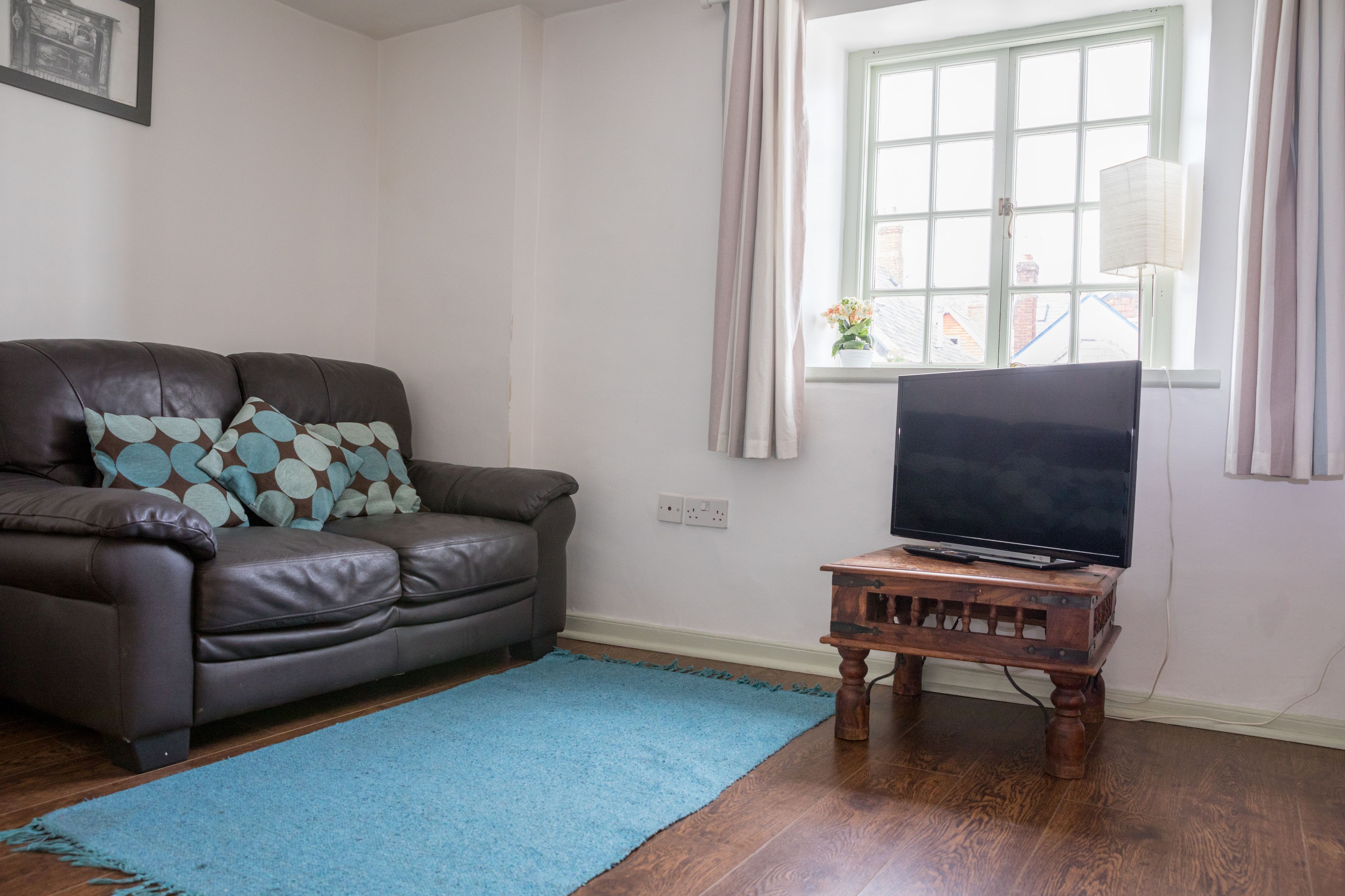 Een woonkamer met een bruine leren bank, een blauw vloerkleed, een houten tv-meubel en een raam met gordijnen.