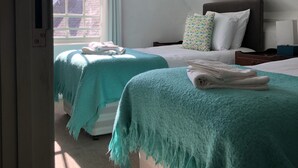Ein Schlafzimmer mit zwei Betten, jeweils mit einer türkisblauen Decke, einem Fenster mit Vorhängen und Blick auf Gebäude.