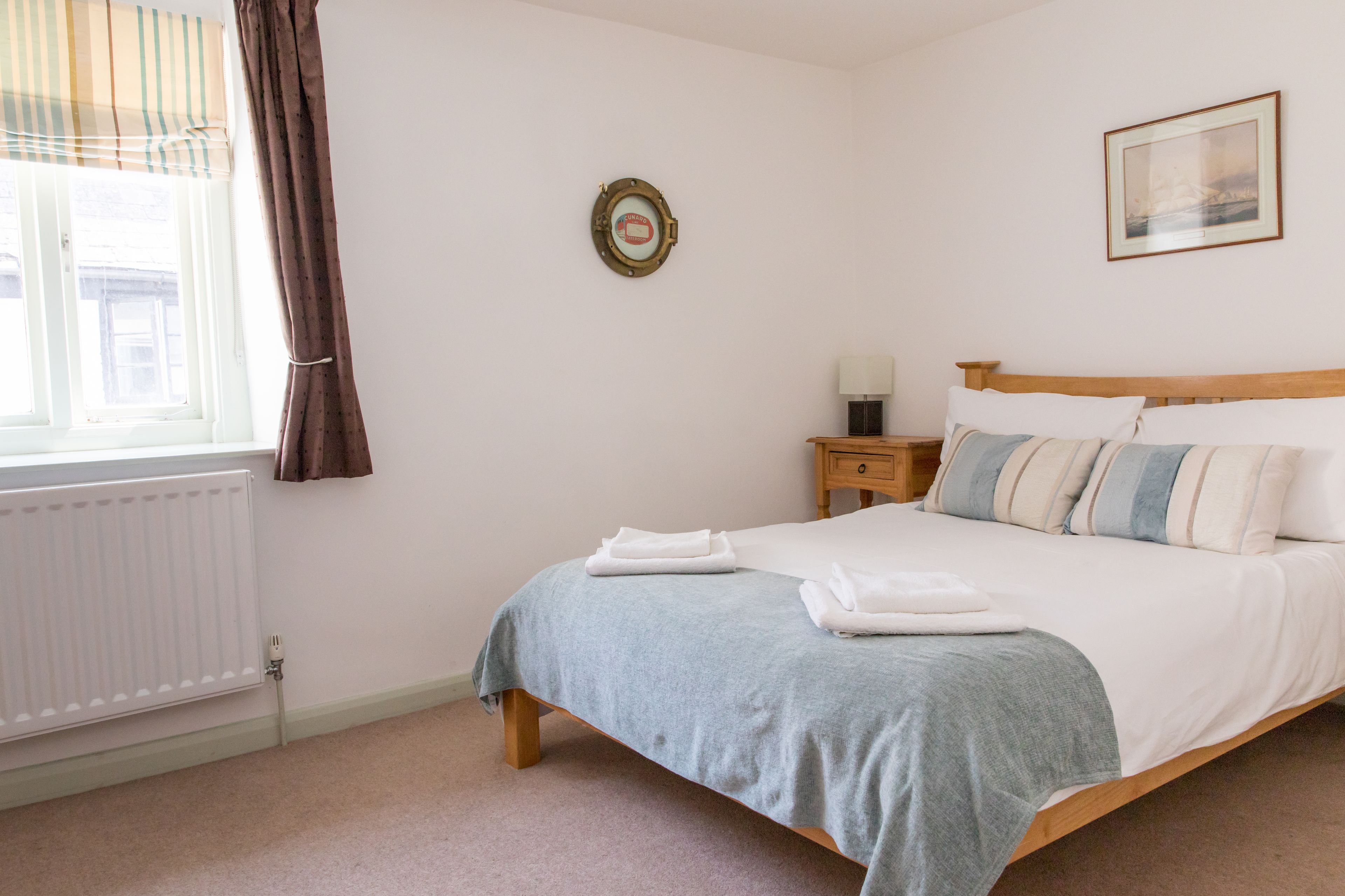 A neatly made bed with a blue blanket and white pillows, a wooden nightstand with a lamp, a window with curtains, and a framed picture on the wall.