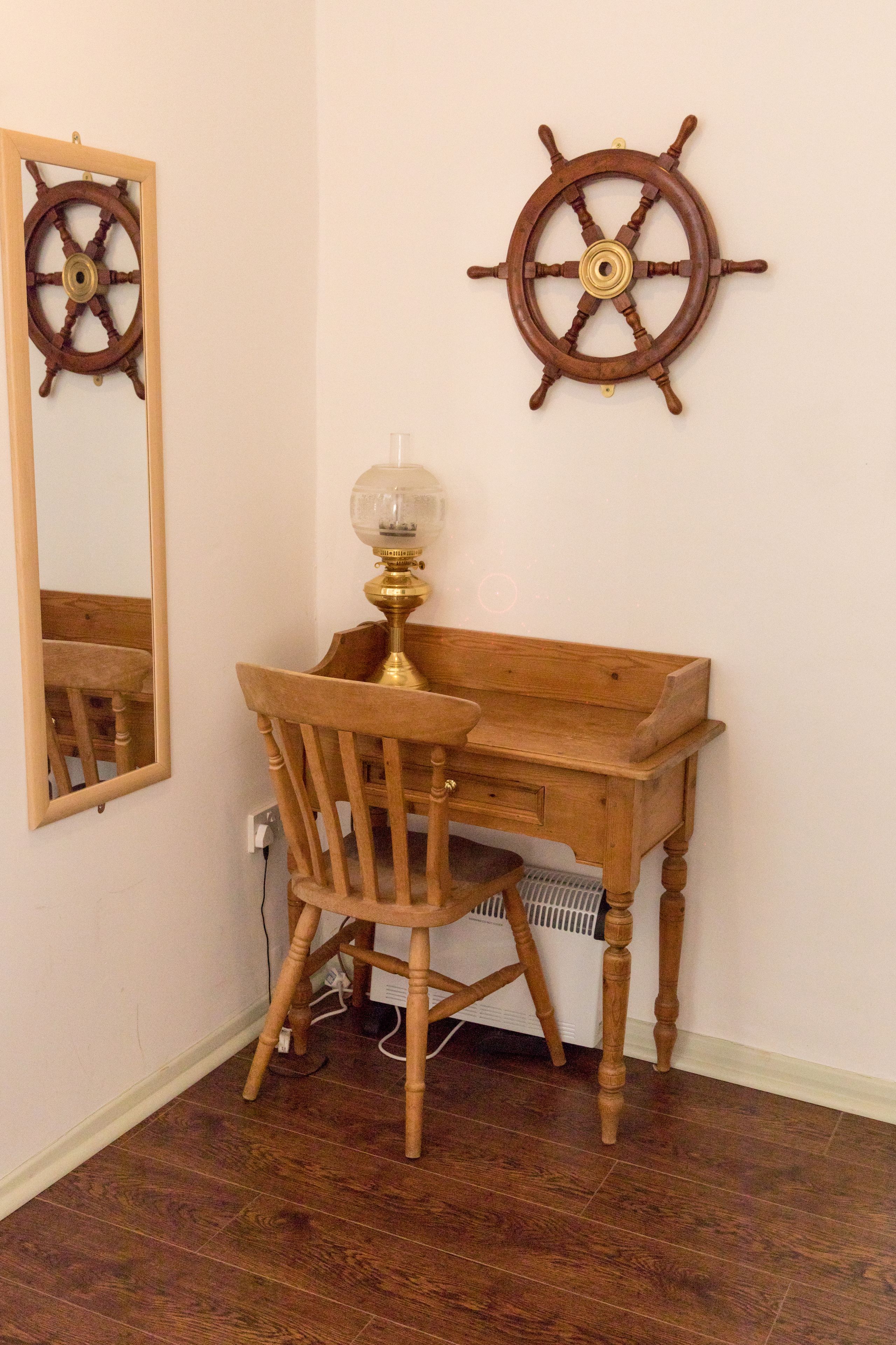 A wooden desk with a chair, a wall-mounted ship's wheel, and a mirror reflecting the room.