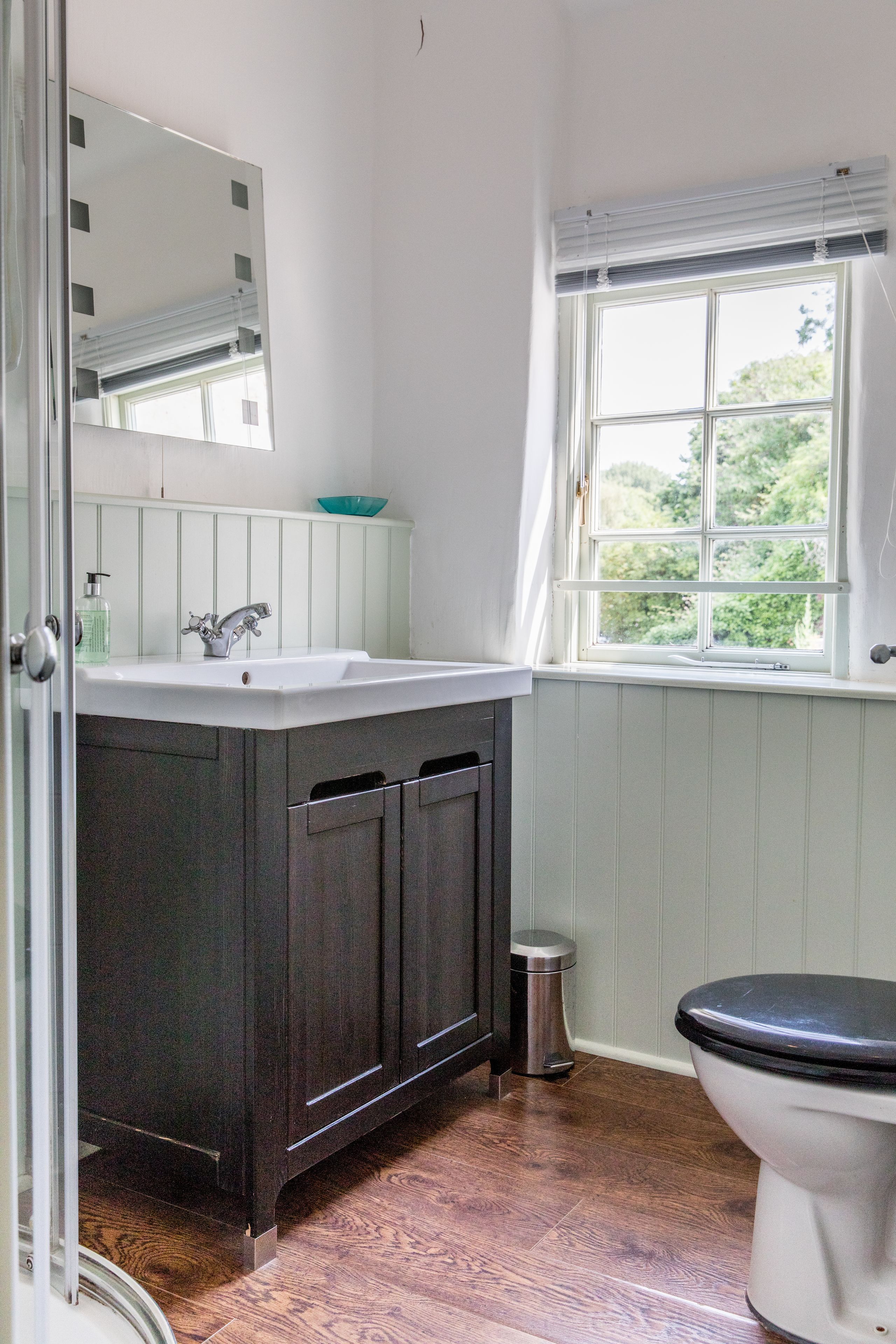Un baño con un lavabo blanco, armarios de madera oscura, un inodoro blanco y una ventana con vistas a zonas verdes.