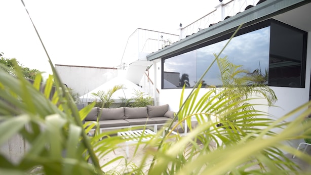 Uma área de estar moderna ao ar livre, com um sofá branco e uma mesa de vidro, rodeada por plantas verdes exuberantes.
