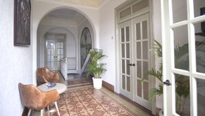 Um corredor com piso estampado, uma poltrona de couro marrom, uma mesa branca com livros, uma planta em vaso e uma porta com painéis de vidro.