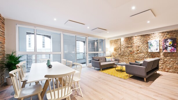 A spacious living area with a large white table and chairs, a grey sofa, and a yellow patterned rug. There is a brick wall with artwork and a large window providing natural light. - Temple Bar Crowne Square Apartments (Dublin)