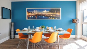 A dining area with a blue wall, a framed cityscape picture, and a table set for four with orange chairs. - Temple Bar Crowne Square Apartments (Dublin)