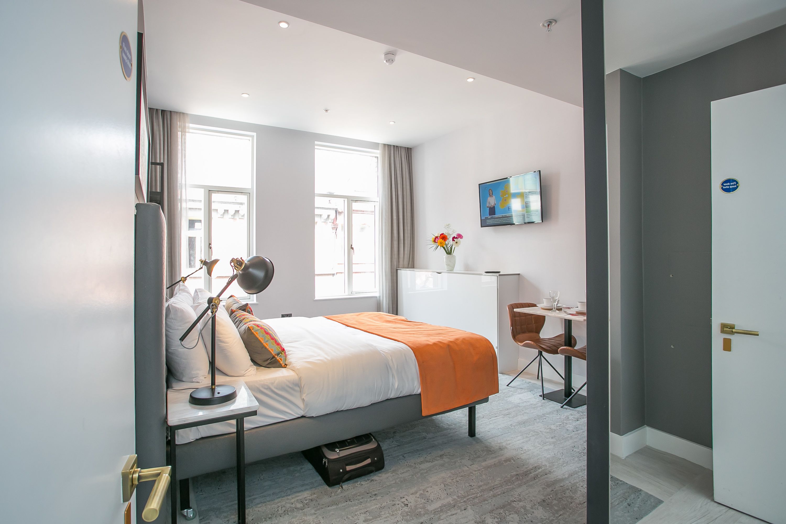 A modern hotel room with a bed, bedside table, lamp, desk, chair, and a TV on the wall.
